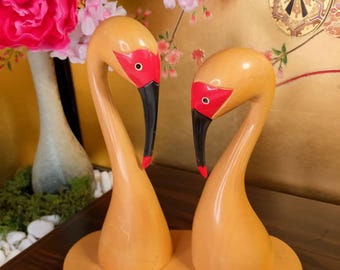 May include: A pair of wooden swan figurines with a light brown finish. The swans have red accents on their faces and black beaks. They are mounted on a matching wooden base, creating a decorative display piece.