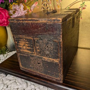 May include: A rectangular dark brown wooden chest featuring a hinged lid, a small upper drawer with a ring pull, and two larger drawers below, each secured with weathered metal latch plates and keyholes, showing signs of aged wood wear.
