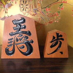 May include: Two wooden Japanese Shogi pieces with black calligraphy. The larger piece features the character for "King", while the smaller piece displays the character for "Pawn". Both pieces are set against a gold background.