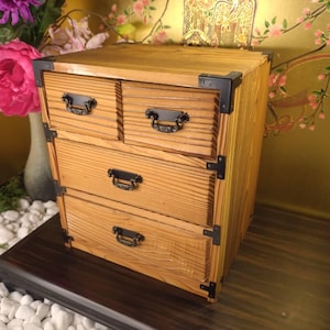 May include: A wooden chest of drawers with four drawers and black metal hardware. The chest is made of light-colored wood and has a natural wood grain finish. The chest is sitting on a dark brown surface.