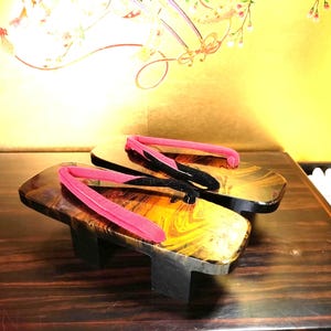 Puede incluir: Un par de sandalias geta japonesas tradicionales. Las plataformas de madera tienen un acabado oscuro y brillante con detalles dorados y amarillos. Las correas son de color rosa brillante y negro. Las sandalias se exhiben sobre una superficie de madera oscura.