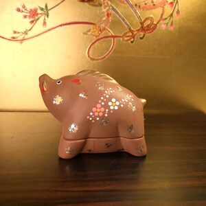 May include: A brown ceramic figurine of a boar with decorative floral and silver accents. The boar has red accents on its snout and ears. The figurine is on a dark wooden surface with a gold background.