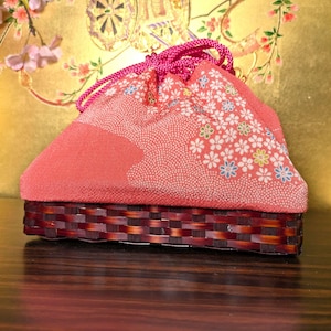 May include: A drawstring pouch featuring a coral-colored fabric top with a white floral pattern and a stippled texture. The base is constructed from a dark brown woven material with a basket-weave design, secured by bright pink braided cords.