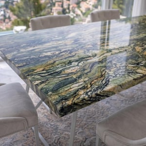 Op de afbeelding: Een rechthoekige eettafel met een gepolijst groen, zwart en goud marmeren blad met een natuurlijk wervelend patroon. De tafel wordt ondersteund door een metalen frame en is omringd door vier lichtbeige gestoffeerde stoelen met gewatteerde rugleuningen.