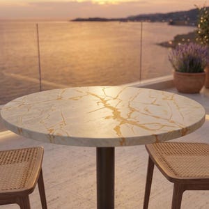 Op de afbeelding: Een ronde tafel met een wit oppervlak met gouden aders, ondersteund door een centrale donkere voet, vergezeld door twee stoelen met lichthouten frames en rugleuningen en zittingen van gevlochten riet, geplaatst op een balkon.