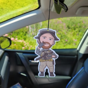 May include: A cartoon air freshener featuring a soldier with a beard, wearing a hat, and holding a rifle. The air freshener is hanging in a car, with a green background visible through the window.