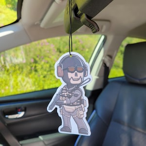 May include: A car air freshener featuring a cartoon illustration of a character in a tactical outfit with a skull face, wearing a helmet and holding a gun. The air freshener is hanging from a black string in a car.
