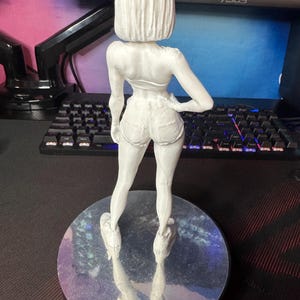May include: A white 3D-printed figurine of a woman in shorts and a top, standing on a round, reflective base. The figure is facing away from the camera, with one hand on her hip. The background includes a computer monitor and keyboard.