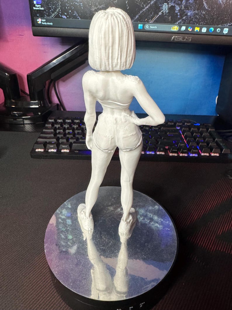 May include: A white 3D-printed figurine of a woman in shorts and a top, standing on a round, reflective base. The figure is facing away from the camera, with one hand on her hip. The background includes a computer monitor and keyboard.