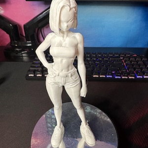 May include: A white 3D-printed figurine of a female character, posed with one hand on her hip. She wears a tube top, shorts, and sneakers. The figure is standing on a reflective, circular base.