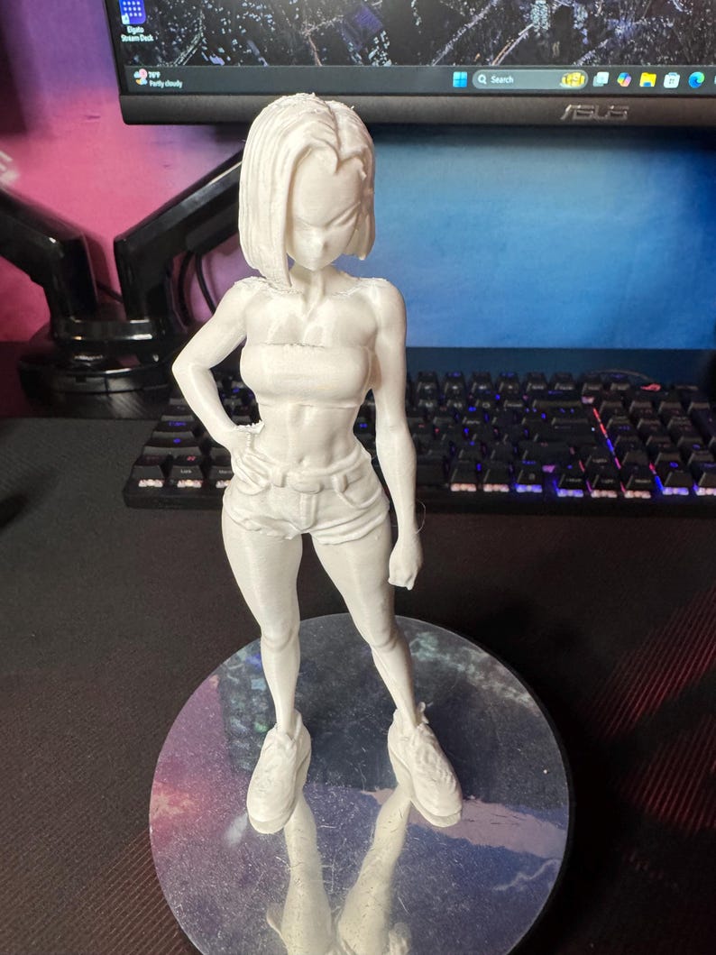 May include: A white 3D-printed figurine of a female character, posed with one hand on her hip. She wears a tube top, shorts, and sneakers. The figure is standing on a reflective, circular base.