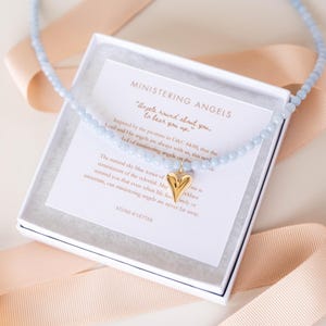 Puede incluir: Un collar de cuentas azul claro con un colgante de corazón dorado dentro de una caja de regalo blanca. La caja contiene una tarjeta con el texto Ministering Angels y una cita. El collar está rodeado por una cinta de color melocotón claro.
