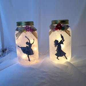 May include: Two glass jars with decorative floral accents and silhouettes of dancers inside. The jars are lit from within, creating a warm glow.