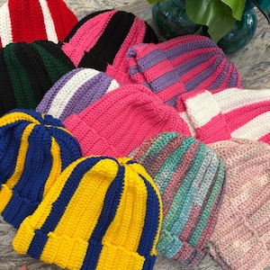 Puede incluir: Una colección de gorros de punto coloridos con varios patrones de rayas. Los sombreros presentan una variedad de colores, incluyendo rojo, rosa, azul, amarillo, verde y morado. Los gorros están hechos de un material suave y texturizado y tienen un dobladillo.