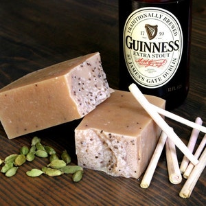 May include: Two bars of handmade soap with a light brown color and a speckled texture. The soap is sitting on a wooden surface with green cardamom pods scattered around it. A bottle of Guinness beer is visible in the background.