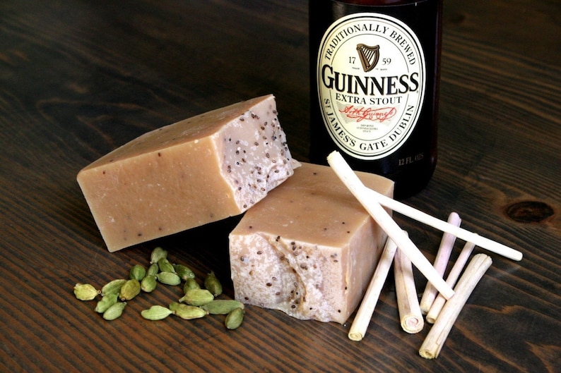 May include: Two bars of handmade soap with a light brown color and a speckled texture. The soap is sitting on a wooden surface with green cardamom pods scattered around it. A bottle of Guinness beer is visible in the background.
