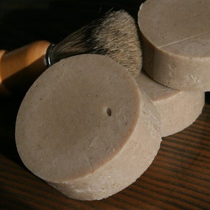 May include: Four round bars of shaving soap stacked on top of each other. The soap is a light brown color and has a rough texture. A shaving brush with a wooden handle is lying next to the soap.