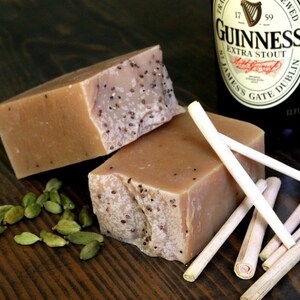 May include: Two bars of brown soap with a speckled texture, a bottle of Guinness beer, and several light brown sticks on a dark wood surface. There are also green cardamom pods scattered around the soap.