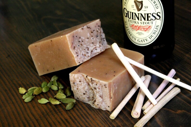 May include: Two bars of brown soap with a speckled texture, a bottle of Guinness beer, and several light brown sticks on a dark wood surface. There are also green cardamom pods scattered around the soap.