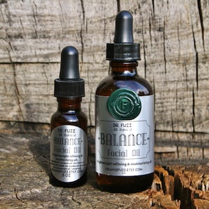 May include: Two glass bottles of facial oil with dropper tops. The bottles are labeled "DR. FUZZ All Natural BALANCE Facial Oil" and have a silver label with green wax seals. The smaller bottle is 0.5 oz and the larger bottle is 1 oz.