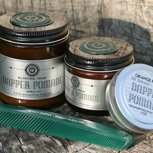 May include: Two amber glass jars of hair pomade with black lids and silver labels that read "Dapper Pomade" and "Orange Fuzz All Natural Vegan Dapper Pomade". A green comb is in the foreground.