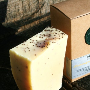 May include: A bar of light yellow soap with black seeds embedded in it. The soap is packaged in a brown box with a green wax seal and the text "ORANGE FUZZ All Natural Handmade Lemon & Chia Seed Soap" printed on the box.