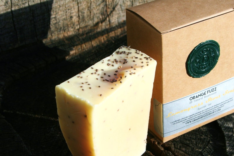 May include: A bar of light yellow soap with black seeds embedded in it. The soap is packaged in a brown box with a green wax seal and the text "ORANGE FUZZ All Natural Handmade Lemon & Chia Seed Soap" printed on the box.