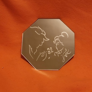 Może przedstawiać: Ośmiokątne lustro z wytrawionym wzorem przedstawiającym sylwetki Bestii i Belli z Pięknej i Bestii. Projekt pokazuje Bestię ofiarowującą różę Belli. Lustro jest umieszczone na pomarańczowym tle.