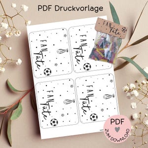 Pode incluir: Uma folha PDF imprimível com quatro etiquetas retangulares contendo o texto "FAN Tüte", ilustrações de bolas de futebol e pequenos pontos. Um saco plástico transparente com doces retangulares coloridos está anexado, com um cabeçalho de cartolina castanha rotulado como "FAN Tüte" e um pequeno gráfico de bola de futebol.