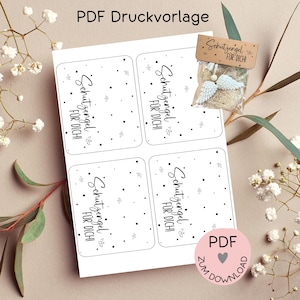 Puede incluir: Cuatro etiquetas de regalo imprimibles con lunares negros y el texto "Schutzengel fur dich!" que se traduce como "¡Ángel de la guarda para ti!" en inglés. Las etiquetas están sobre un fondo blanco. Hay una pequeña bolsa de papel marrón con un amuleto de ala de ángel blanco y el texto "Schutzengel fur dich!" en ella.