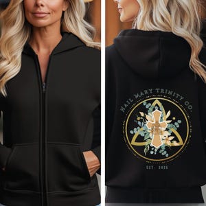May include: Black zip-up hoodie with a gold and green design on the back. The design features a cross inside a circle with the text "Hail Mary Trinity Co. Est. 2025".