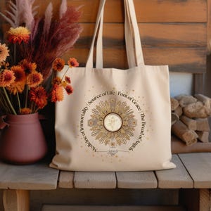 May include: A beige tote bag with a gold and white design featuring a cross and the words "Medicine of Immortality, Source of Life, Font of Grace, The Bread of Angels".
