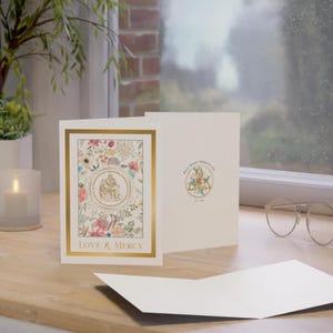 May include: A cream-colored greeting card with a gold border and floral design. The card features the text "LOVE & MERCY" and a circular emblem. The open card reveals a blank interior, with a logo on the back.