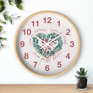 Puede incluir: Un reloj de pared redondo con marco de madera clara. La esfera del reloj es blanca con números y manecillas rojas. Un diseño de corazón floral está en el centro, con las palabras "Love is patient, Love is kind, Love never fails."