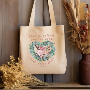 May include: A beige tote bag featuring a floral heart design and the text "Love is patient. Love is kind. Love never fails. 1 COR 13".