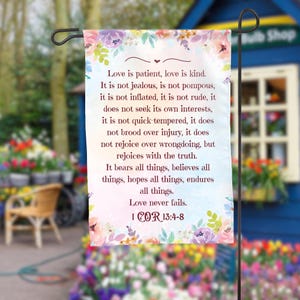 Peut inclure: Un drapeau de jardin avec une bordure florale dans les tons de rose, de violet et de jaune. Le drapeau affiche le texte "Love is patient, love is kind" et des versets de 1 Corinthiens 13:4-8.