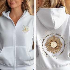 Puede incluir: Sudadera blanca con cremallera y un diseño dorado y blanco en la espalda. El diseño presenta una cruz, un sol radiante y el texto "Medicina de la inmortalidad | Fuente de vida | Fuente de gracia | El pan de los ángeles". La parte delantera de la sudadera tiene un pequeño logotipo con el texto "Hail Mary Trinity Co. Est. 2020".