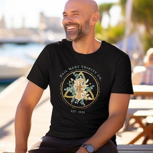 May include: Black t-shirt featuring a gold and floral design with the text "HAIL MARY TRINITY CO." and "EST. 2025". The design includes a cross and a triangle shape. The shirt is a crew neck.