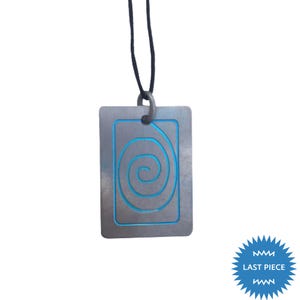 Könnte beinhalten: Rechteckiger Anhänger aus silberfarbenem Metall mit blauem Spiralmuster und blauem Rand, an einer schwarzen Kordel. Der Anhänger hat eine strukturierte Oberfläche. Der Text "LAST PIECE" ist in Sternform blau.