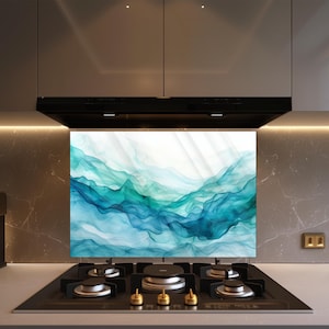 Op de afbeelding: Een keukenachterwand met een abstract ontwerp van vloeiende blauwe en turquoise kleuren, die op aquarel golven lijken. De achterwand is geïnstalleerd boven een zwart kooktoestel met goudkleurige branderknoppen.