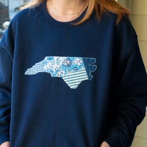 Puede incluir: Sudadera azul marino con un diseño de patchwork del contorno del estado de Carolina del Norte. El parche incorpora varios patrones azules y blancos, incluyendo rayas y estampados florales. La sudadera es de color azul marino liso.