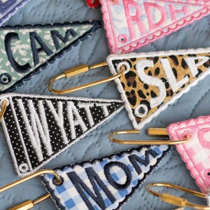May include: Embroidered pennant keyrings in various patterns and colours, including leopard print, gingham, and floral designs. Each keyring features a different name or word, such as "MOM" or "WYATT", with a gold-coloured clasp.