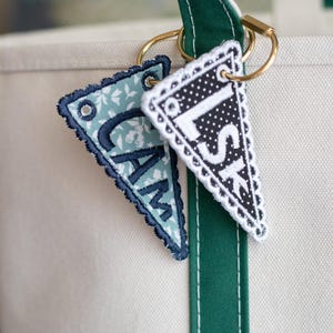 May include: Two personalised pennant keyrings with embroidered letters hang from a gold ring attached to a green and white tote bag strap. One pennant is light blue with white floral print and the letters "CAM". The other is black and white polka dot with the letters "LSK".