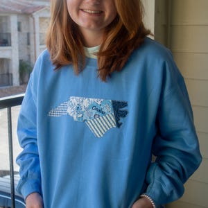 May include: Pale blue sweatshirt featuring a patchwork design of the North Carolina state outline. The patch incorporates floral and striped fabrics, with the word "Carolina" embroidered on it.
