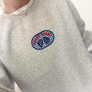 Puede incluir: Una sudadera gris claro con un parche de cuadros azules y blancos. El parche presenta dos ilustraciones de ostras y las palabras "SHUCK YEAH!" en rojo. La sudadera es de estilo casual.