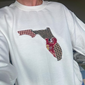Può includere: Felpa bianca girocollo con applicazione a forma di stato della Florida. L'applicazione è realizzata con vari tessuti fantasia, tra cui quadri e righe, e presenta un logo dei Seminoles.