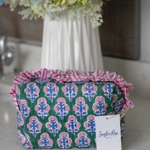 May include: A green cosmetic bag with a floral pattern in pink and blue. The bag has a pink and white striped ruffle trim. A white tag with the text "Saylor Rae" is attached.