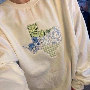 Może przedstawiać: Biała bluza z haftem przedstawiającym stan Teksas w stylu patchwork. Patchwork składa się z różnych tkanin kwiatowych i kratkowych w odcieniach niebieskiego, zielonego i białego.