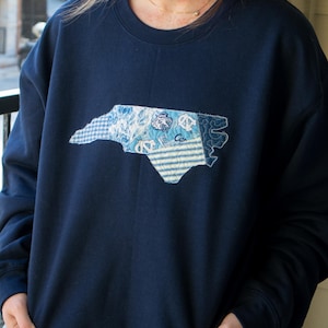 K&ouml;nnte beinhalten: Marineblaues Sweatshirt mit einem Patchwork-Design des Umrisses des Bundesstaates North Carolina. Der Patch enth&auml;lt verschiedene blaue Muster und das Wort "Car". Ein l&auml;ssiges, bequemes Oberteil f&uuml;r den Alltag.