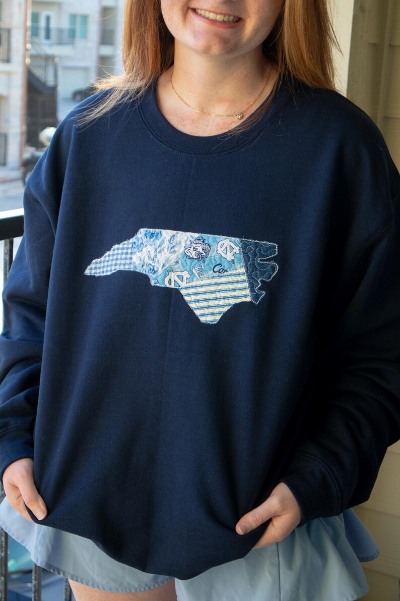 K&ouml;nnte beinhalten: Marineblaues Sweatshirt mit einem Patchwork-Design des Umrisses des Bundesstaates North Carolina. Der Patch enth&auml;lt verschiedene blaue Muster und das Wort "Car". Ein l&auml;ssiges, bequemes Oberteil f&uuml;r den Alltag.